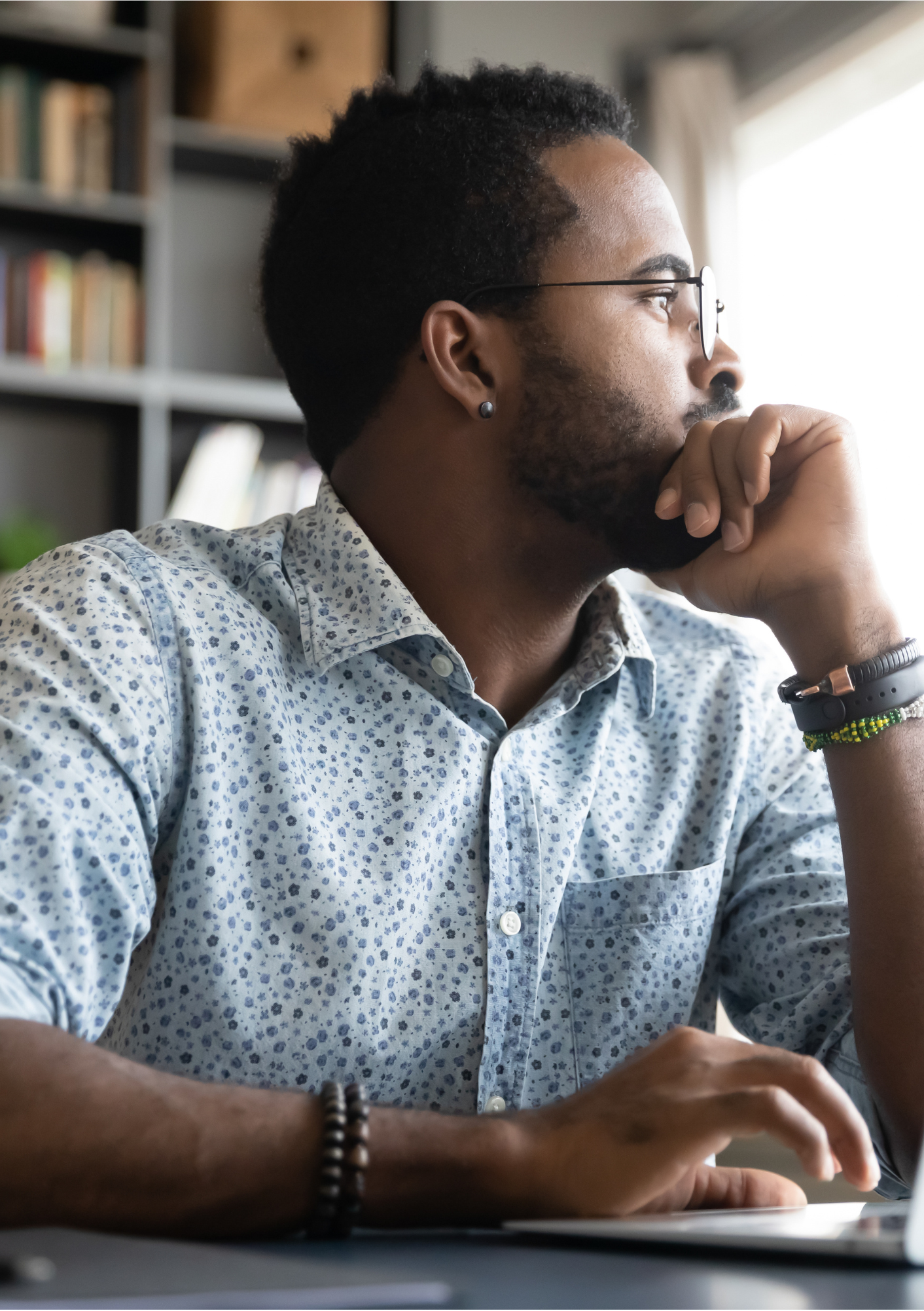 Man sitting at a desk with a laptop, looking thoughtful with a bookshelf in the background.