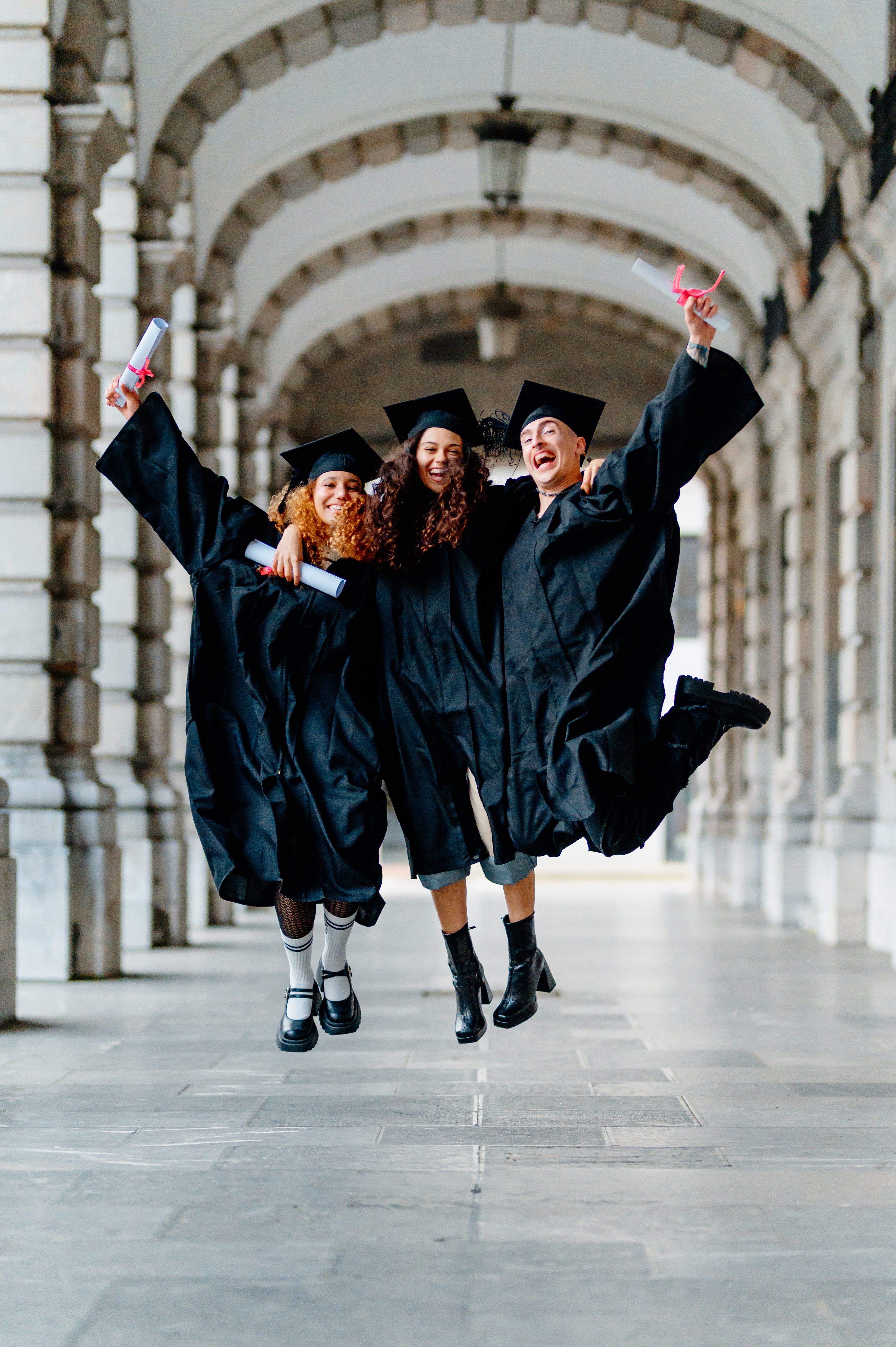 Three graduates in black gowns and caps jumping in a celebratory manner in an architectural setting.