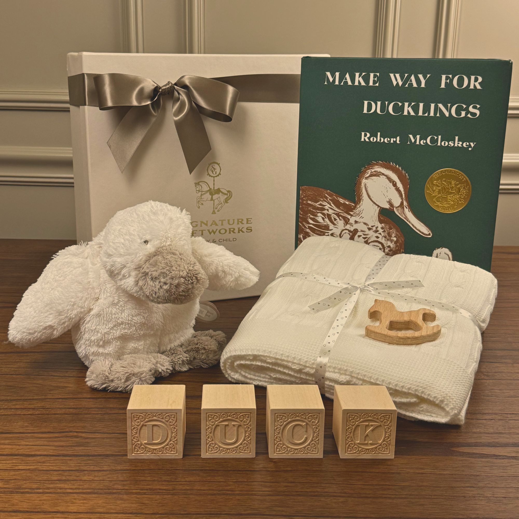 Baby gift set with plush toy, book, blocks, and blanket on a wooden surface.