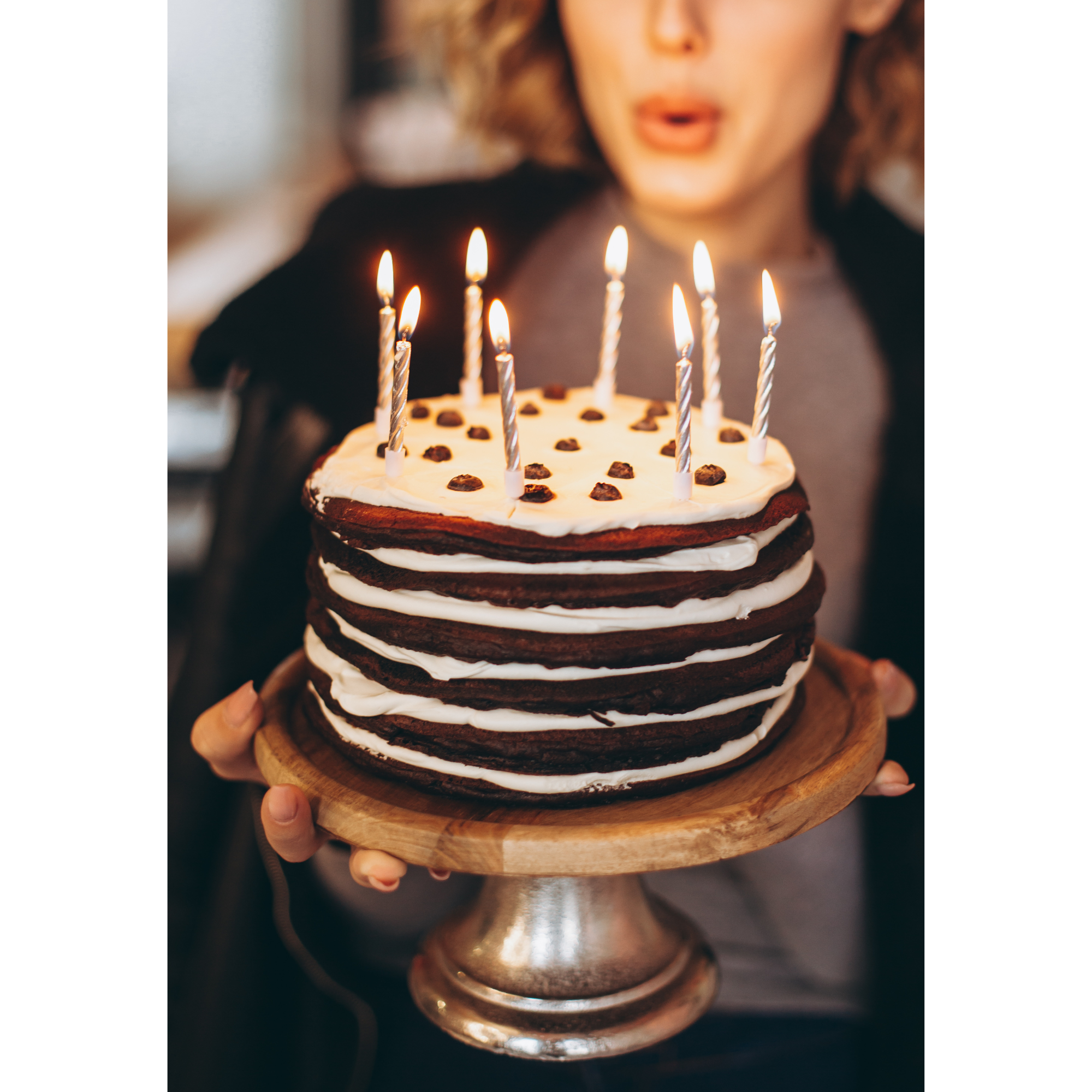 Person holding a layered chocolate cake with lit candles on a wooden stand.